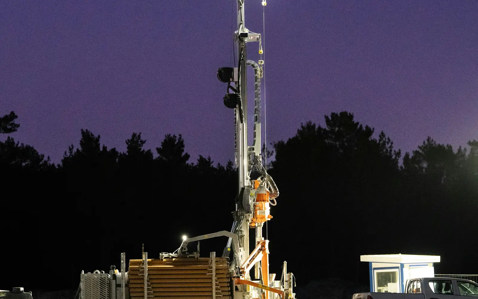 A borehole drilling rig working under floodlights at dusk, casing pipe stacked beside it