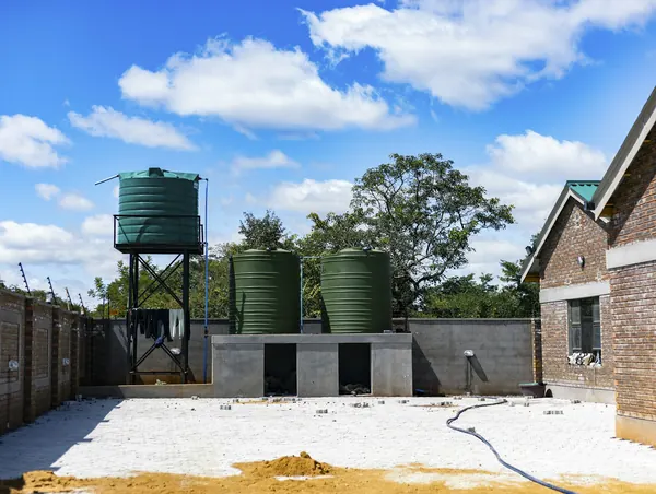 Green water tanks on a steel stand and a concrete base beside a brick house